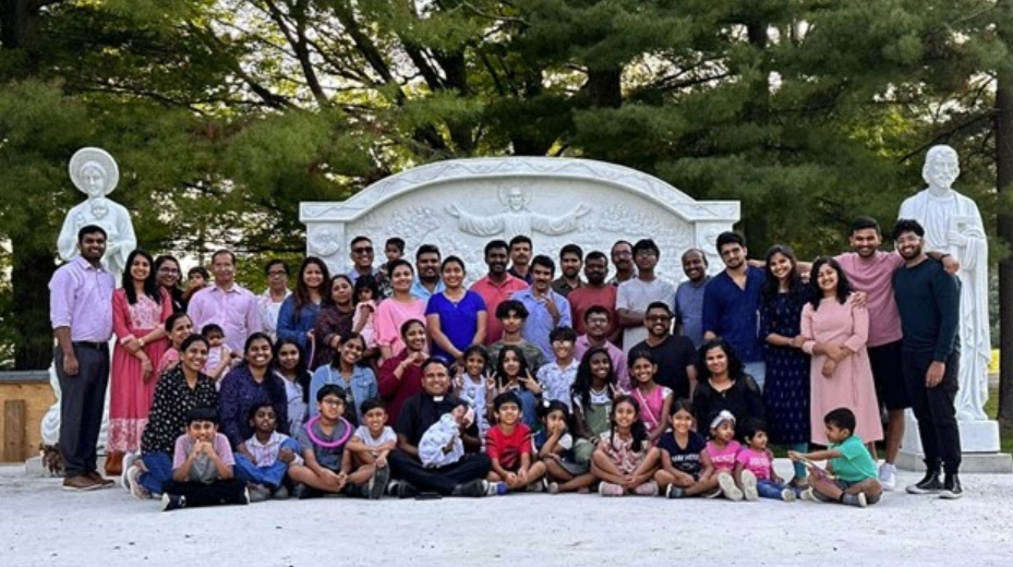 Large community group gathered at a sacred pilgrimage site with white stone religious monument and statues surrounded by lush greenery